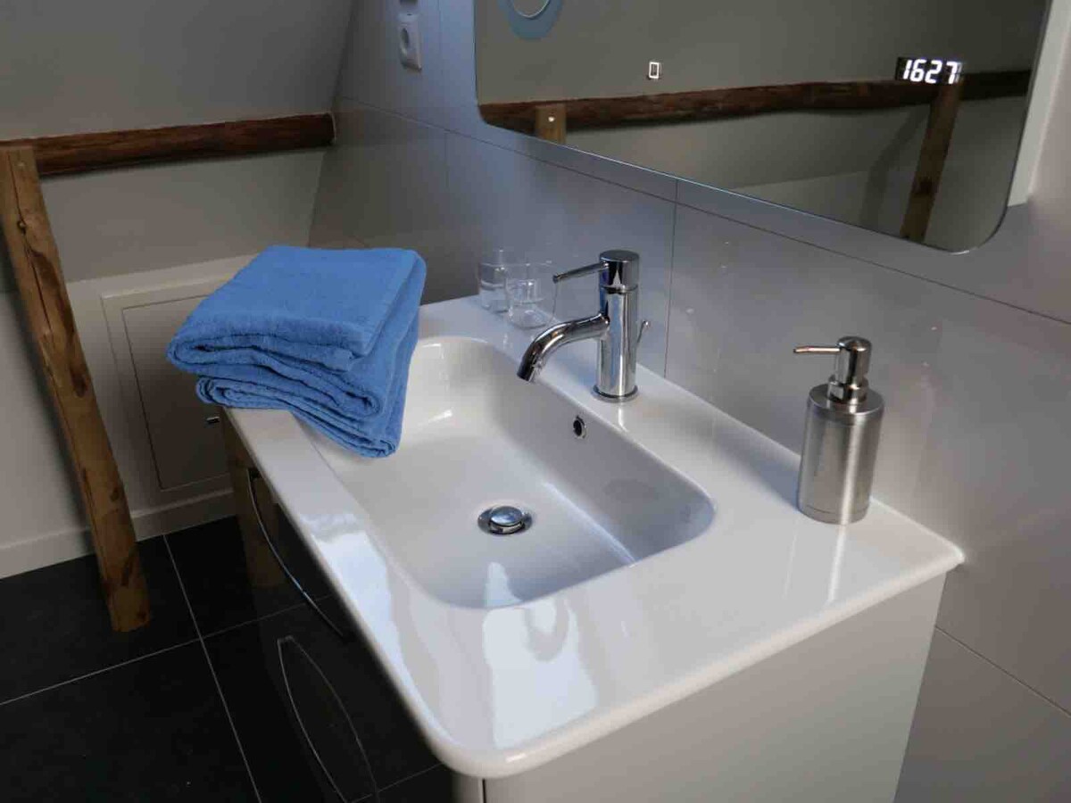 A clean and modern bathroom sink area is displayed. A white sink is featured with a polished faucet, accompanied by a glass soap dispenser and a set of blue towels neatly arranged. The reflection in the mirror highlights the room's simplicity and contemporary design.