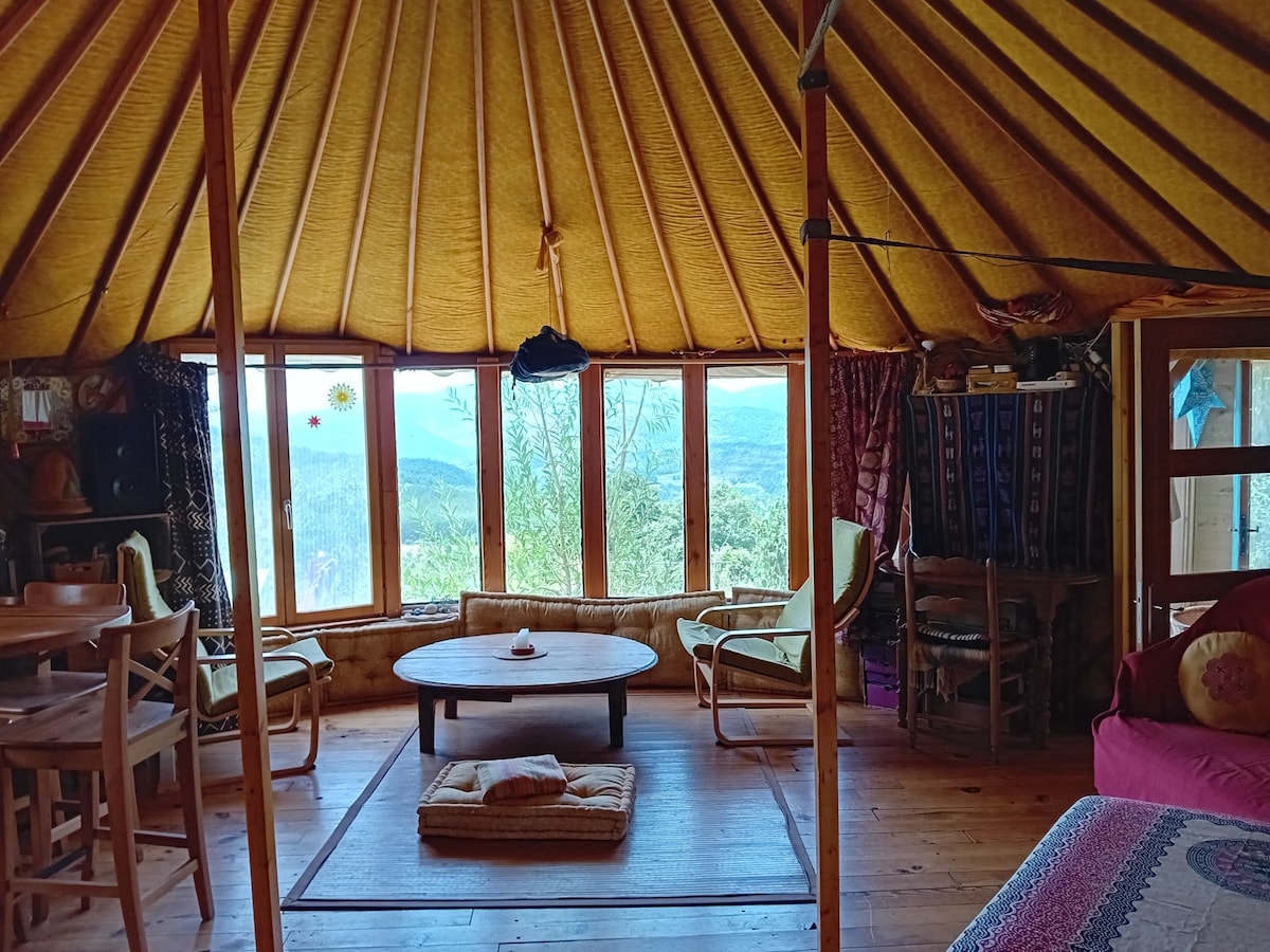 The interior of the yurt features warm-colored walls and a circular ceiling that draws the eye. A low wooden table is surrounded by comfortable seating, with large windows framing a panoramic view of the mountains and village beyond.