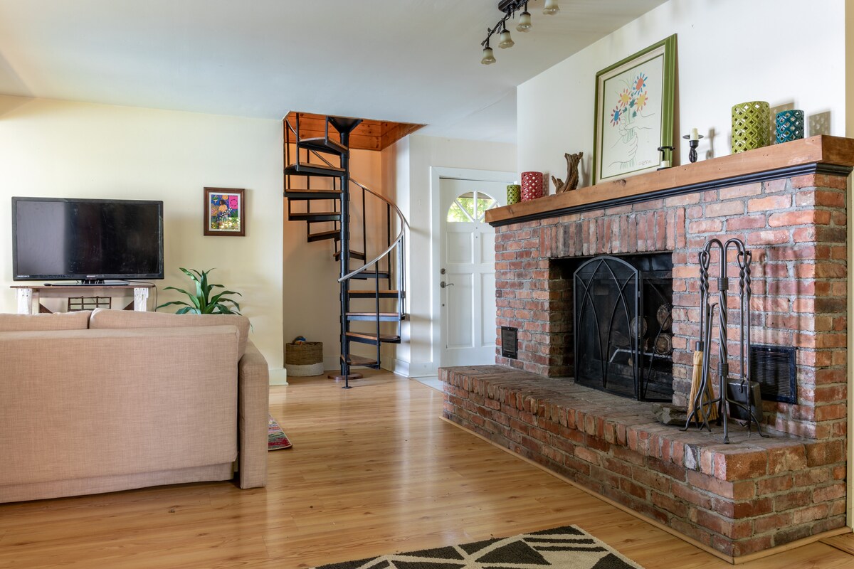 The living area features a brick fireplace with a simple black screen, complemented by a light-colored sofa and a large TV. A spiral staircase leads to the upper floor, while natural light enters through an adjacent doorway. Decorative items are arranged on the mantel.