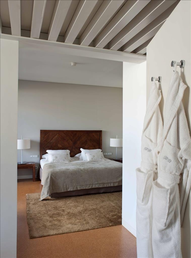 The entrance opens to a spacious room featuring a wooden bed with soft linens. Two bedside lamps frame the bed, while plush brown rugs add warmth to the cork floor. Two bathrobes hang on hooks near the entrance, highlighting the room’s comfort.