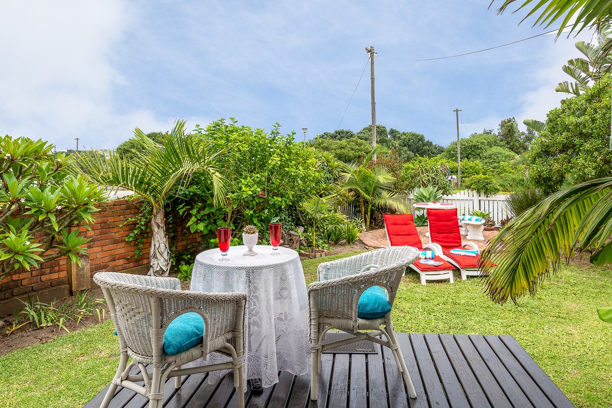 A wooden terrace features a round table with two wicker chairs, accented by turquoise cushions. The setting includes a view of a lush garden with recliners and vibrant red loungers, creating an inviting outdoor space for relaxation.