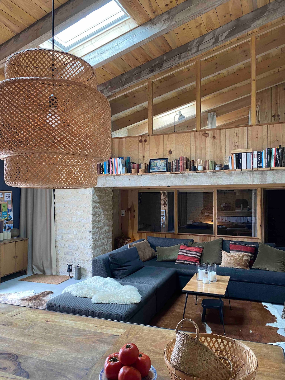 An airy living space features a sectional sofa adorned with pillows, complemented by a wooden coffee table. A woven pendant light hangs above, and bookshelves line the upper balcony. Natural light filters through skylights, illuminating the warm wooden beams and stone walls.