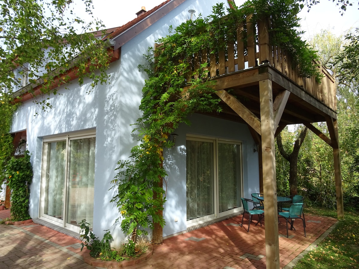 The exterior of the studio gîte features large windows framed by lush greenery. A cozy seating area with two chairs is positioned near the entrance. Above, a small deck is supported by wooden beams, offering additional outdoor space surrounded by gardens.