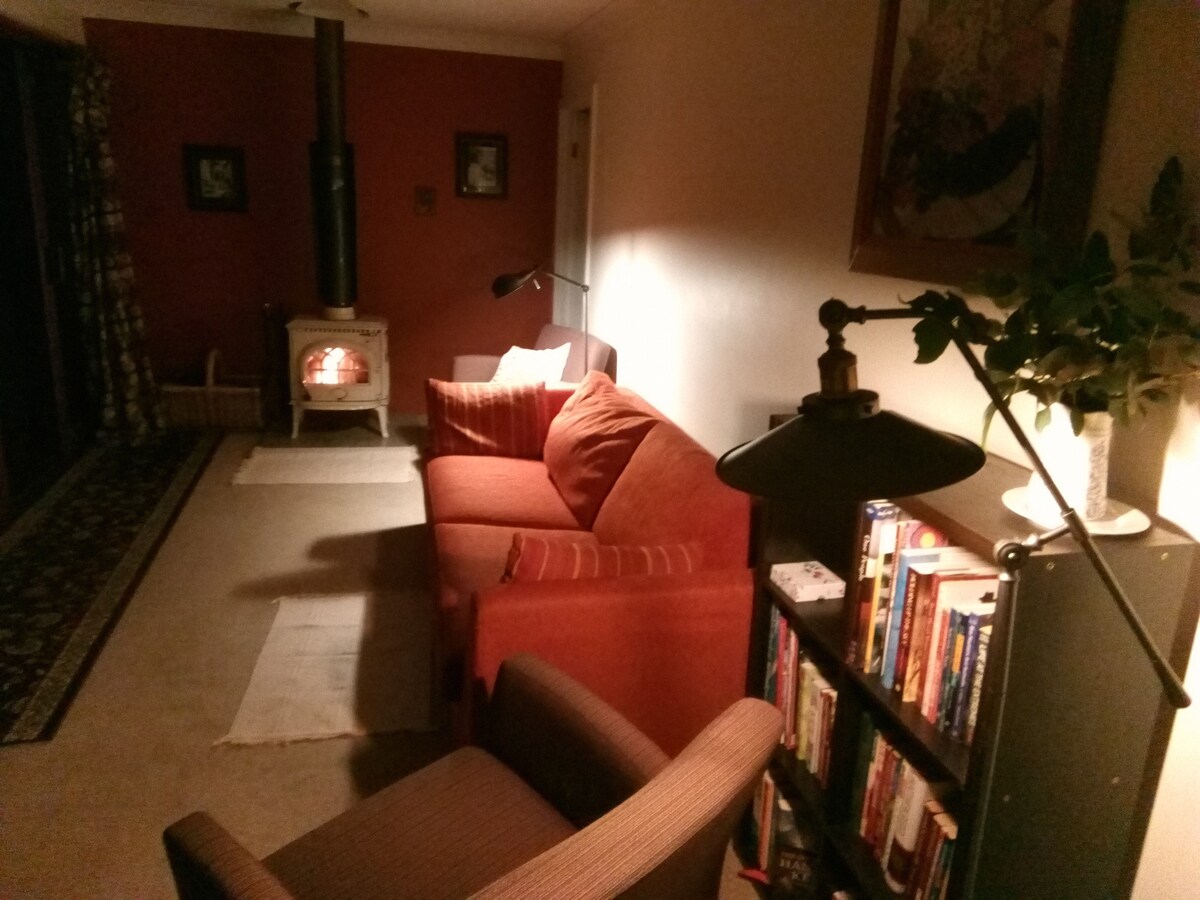 A living area is illuminated by a warm glow from a wood-burning stove, adding to the cozy ambiance. A pair of comfortable chairs and a bright red sofa, decorated with cushions, are positioned near a bookshelf filled with books. Soft lighting creates an inviting space.