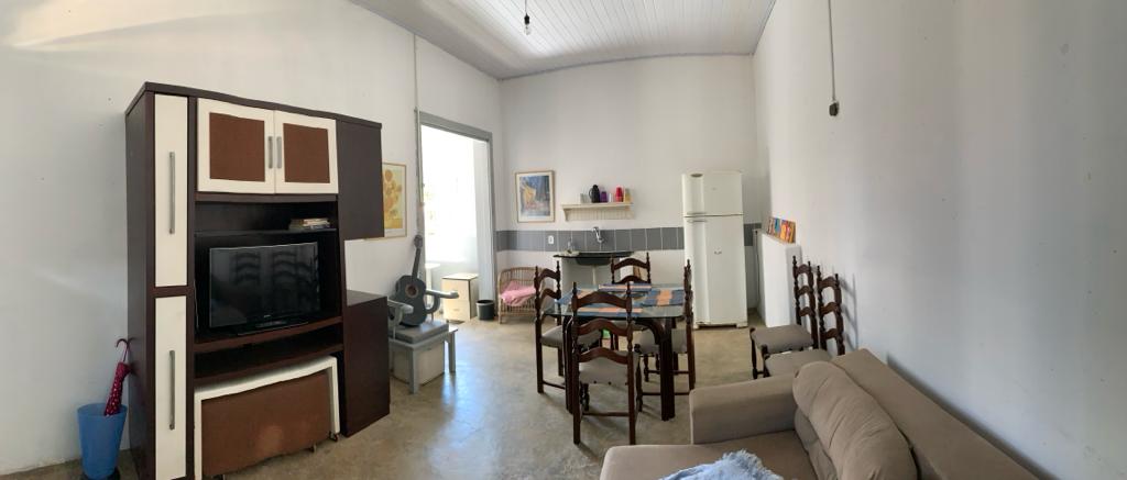 A simple living area features a comfortable sofa and a glass dining table surrounded by wooden chairs. A television unit stands against the wall, while a refrigerator and additional seating are visible. Natural light enters through a window, illuminating the space.