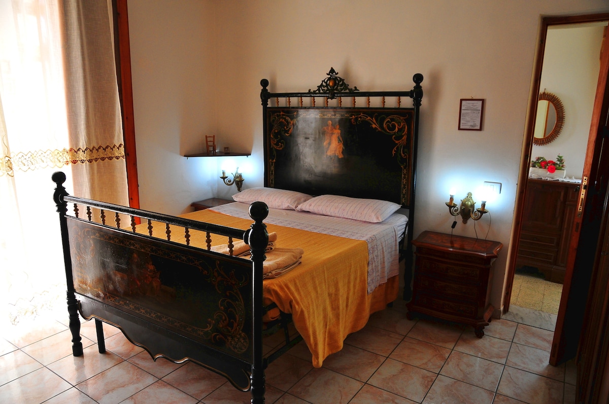 An antique French bed, dating back to 1901, is centered in the room, complemented by soft linens. Ambient lighting from wall sconces adds a gentle glow. A doorway leads to a shared bathroom, while tiles underfoot provide a classic touch.