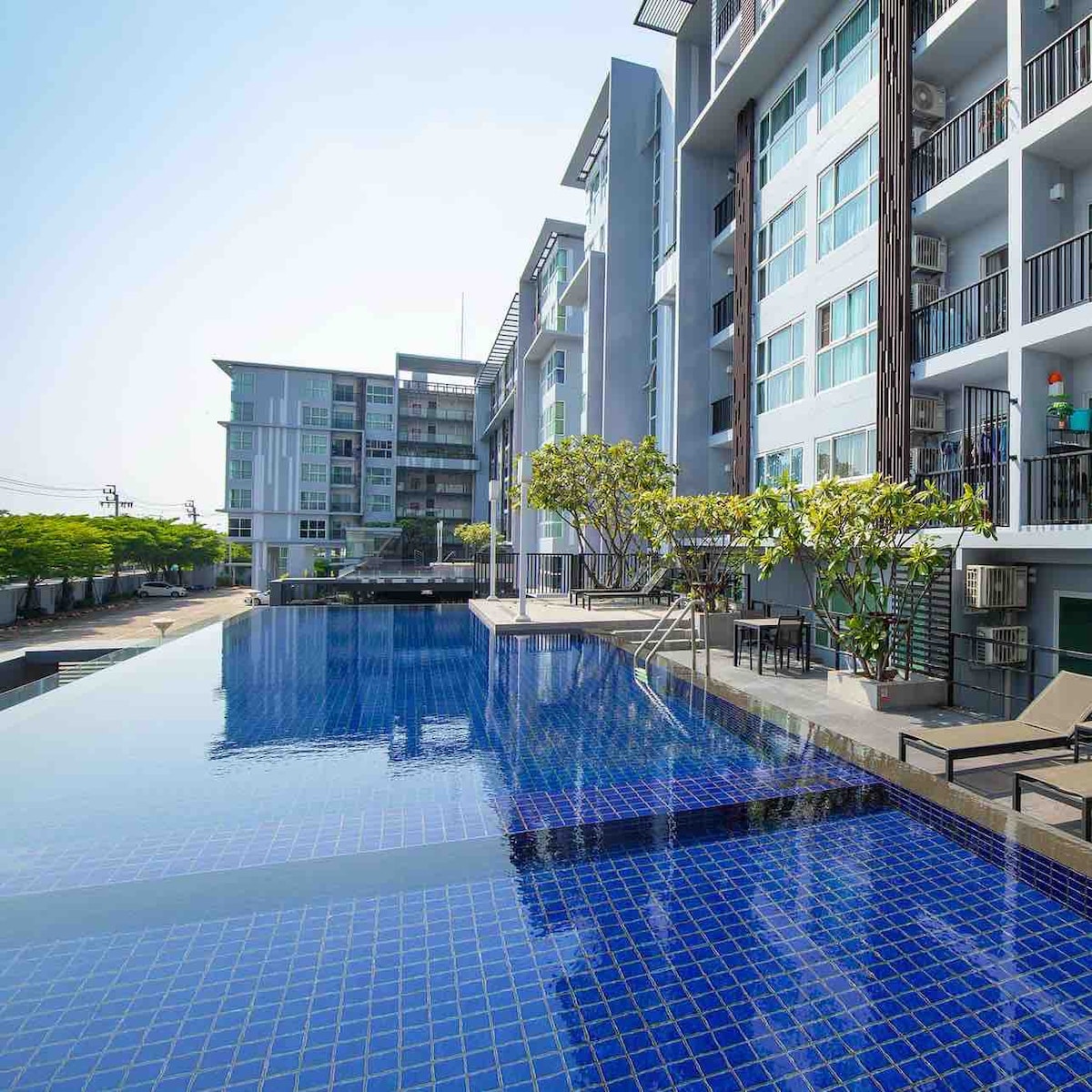 An infinity pool is visible, reflecting the clear sky and surrounded by well-maintained greenery. The condominium buildings stand in the background, showcasing modern architecture with large windows. Lounge chairs are placed along the poolside, offering a serene space for relaxation.