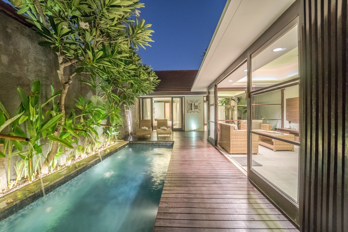 A private pool surrounded by lush greenery is visible, with wooden decking leading towards an indoor living area. Large glass doors create a seamless transition between the outdoor space and the comfortable seating inside.