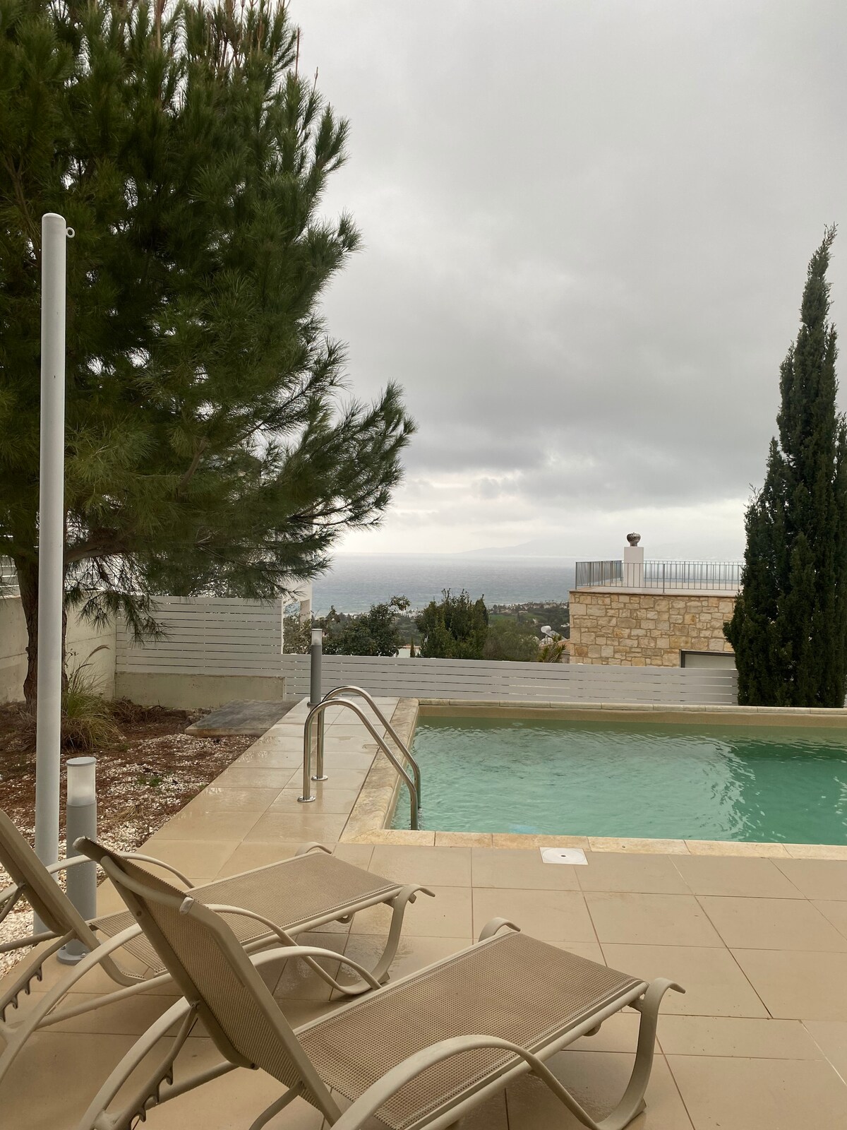 An outdoor area is shown with two lounge chairs positioned beside a clear swimming pool. Lush trees are visible in the background, while distant hills can be seen under a cloudy sky. The serene setting offers a sense of privacy and relaxation.
