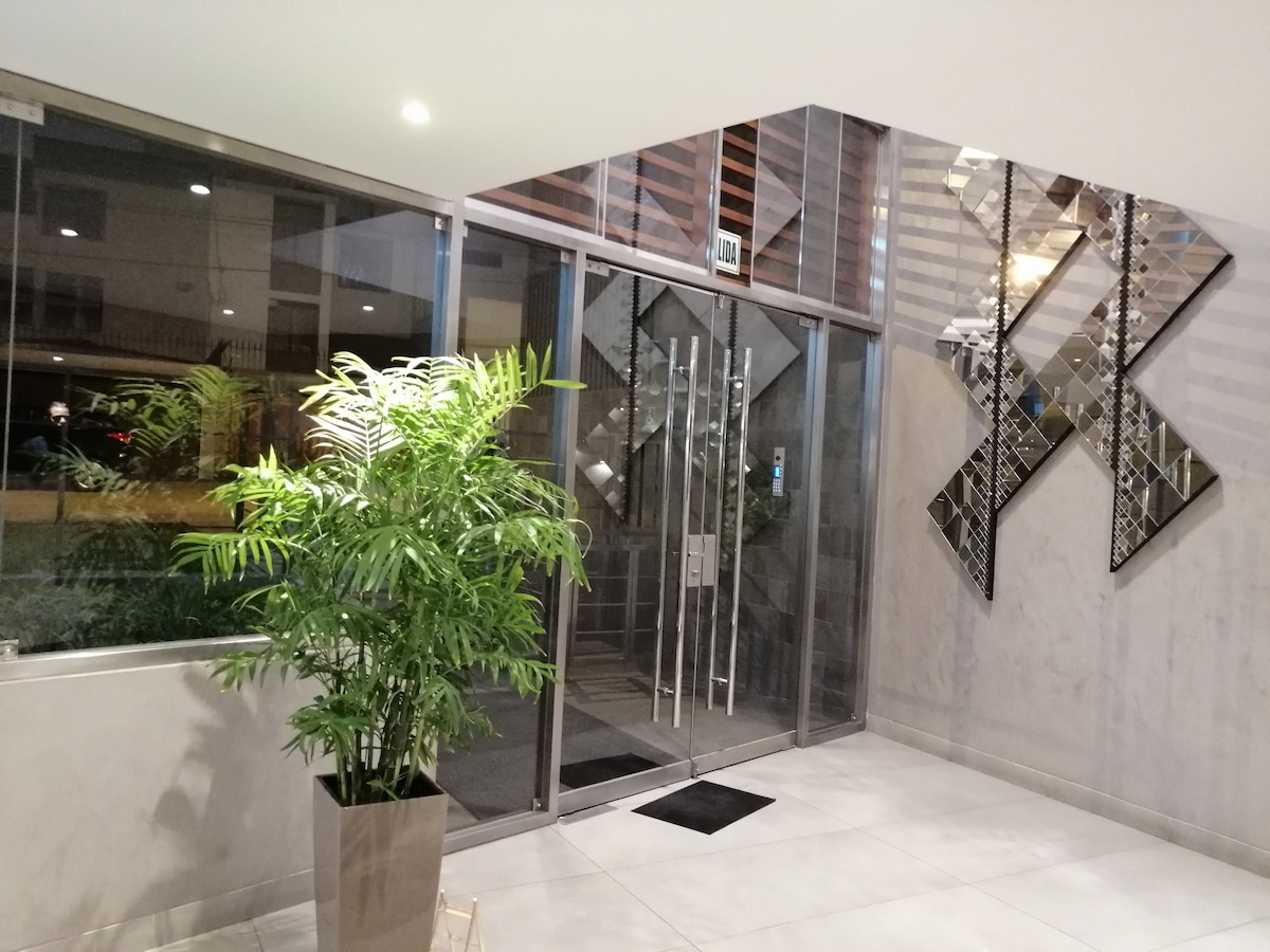 A modern entrance area features a large glass door framed with mirrored accents. A potted plant adds greenery to the space, which is illuminated by ambient lighting. The polished floor complements the contemporary design of the lobby.