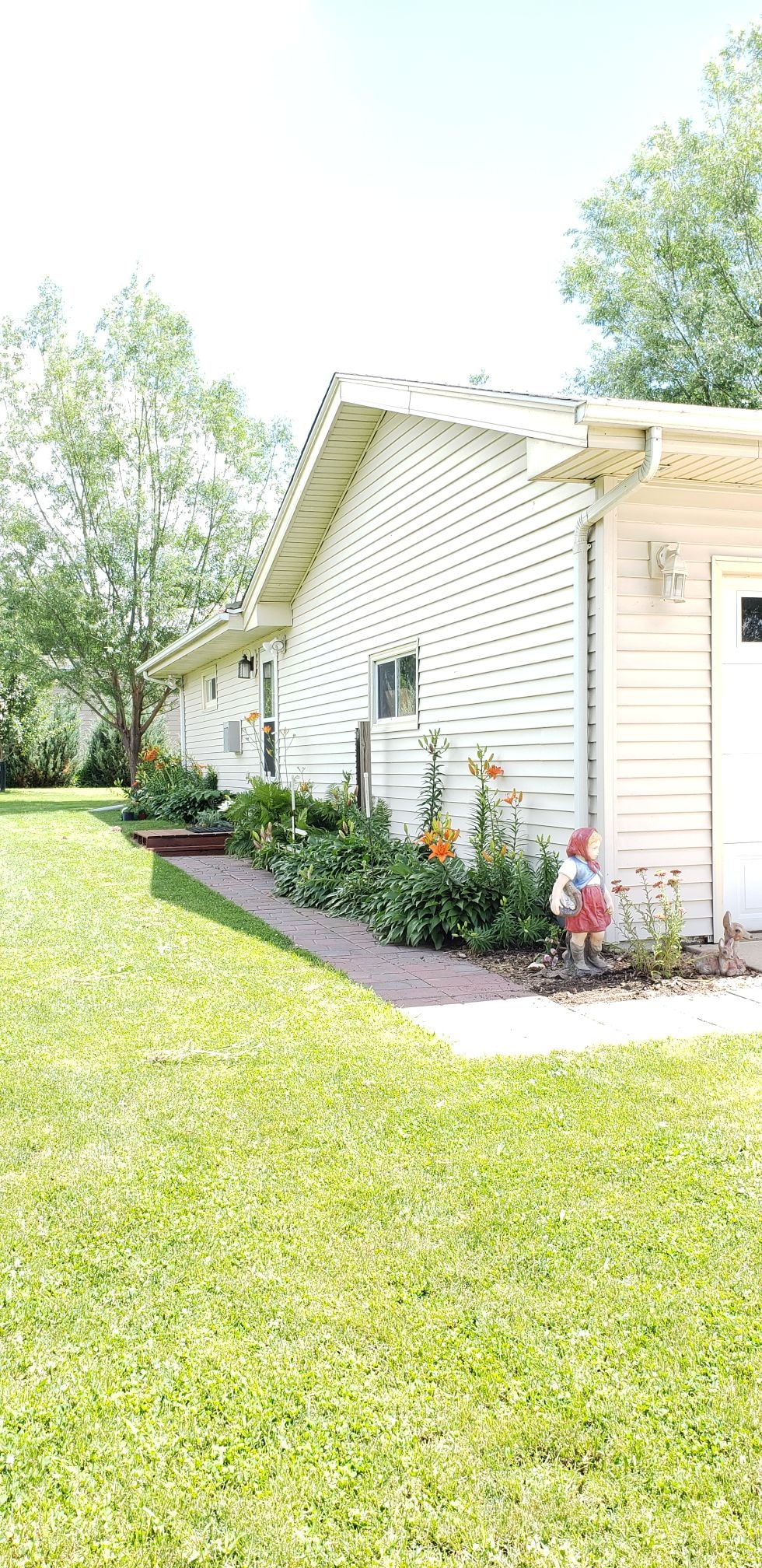 A well-maintained exterior of the private studio apartment is visible, showcasing light-colored siding and a landscaped garden. Lush green grass surrounds the pathway, leading to the entrance. Decorative elements, including a small statue, add character to the serene outdoor space.
