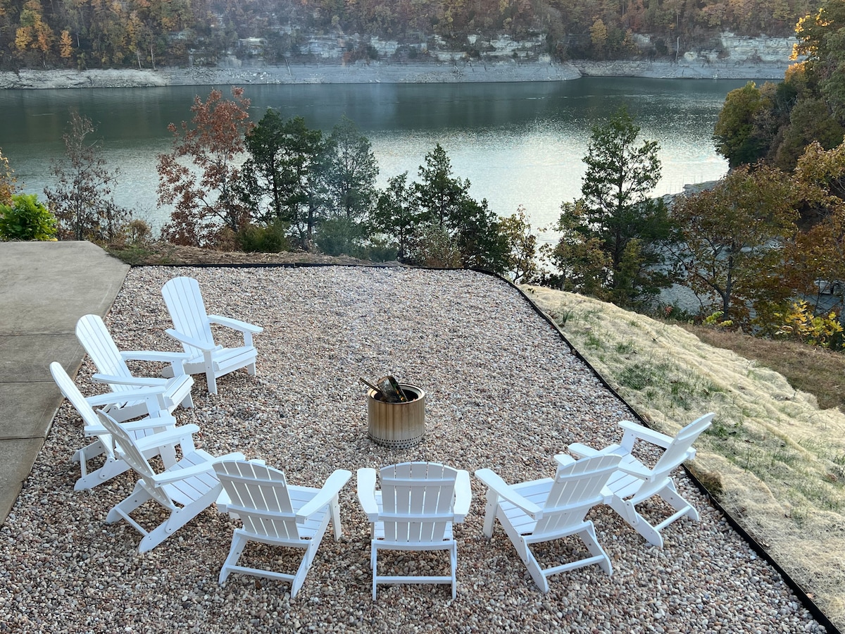 A gathering area features white Adirondack chairs arranged in a circular formation around a central fire pit. The space overlooks a serene body of water with trees in autumn hues surrounding the scene, creating a peaceful outdoor setting.