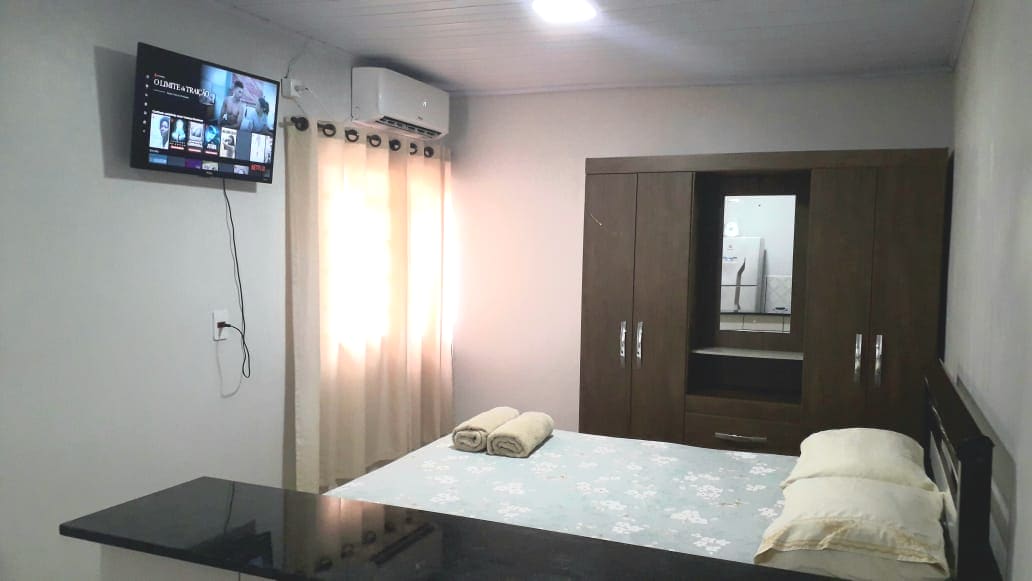 A comfortable sleeping area features a spacious bed with soft pillows and neatly folded towels. A flat-screen TV is mounted on the wall, and natural light filters through sheer curtains, illuminating the light-colored walls. A dark wood wardrobe offers additional storage.