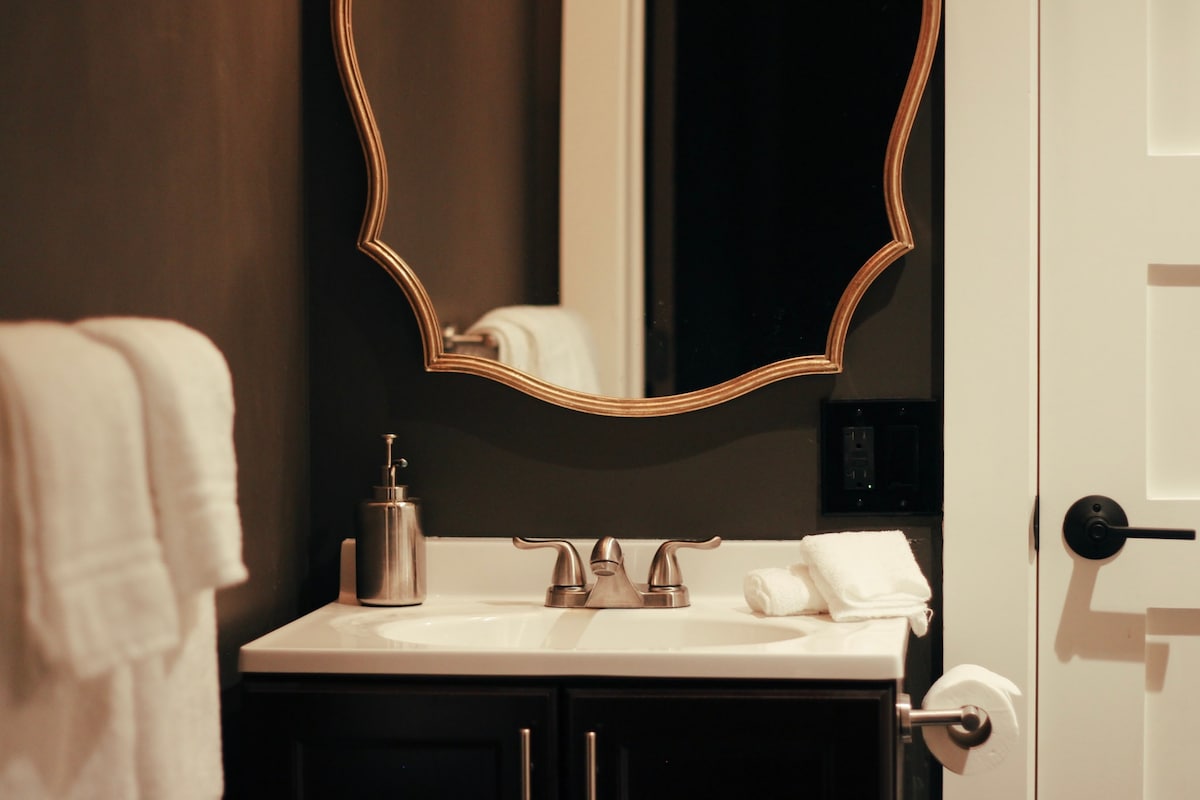 A modern bathroom features a sleek, dark wall and a white countertop with a stylish sink. A decorative mirror with a gold frame hangs above the sink. Fresh white towels are neatly displayed, while a bottle and a folded towel are placed beside the faucet.