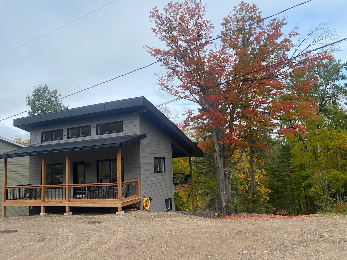 A modern chalet is set against a backdrop of vibrant autumn foliage. The structure features a covered porch that extends across the front, with large windows allowing natural light to fill the interior. Surrounding trees showcase shades of red and orange.