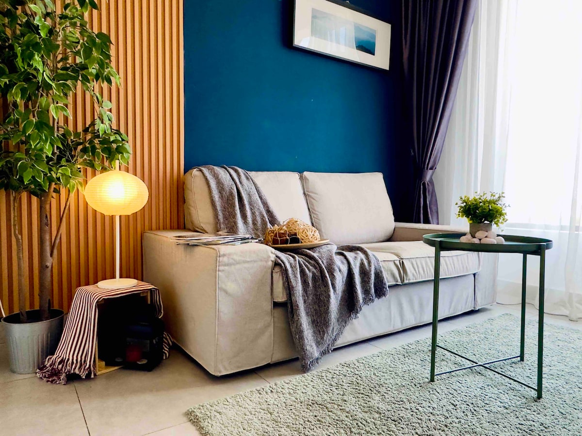 A comfortable seating area features a light-colored sofa draped with a soft blanket. A small green table sits beside the sofa, accompanied by a decorative plant. The backdrop includes a textured blue wall, enhancing the cozy atmosphere with soft lighting from a nearby lamp.
