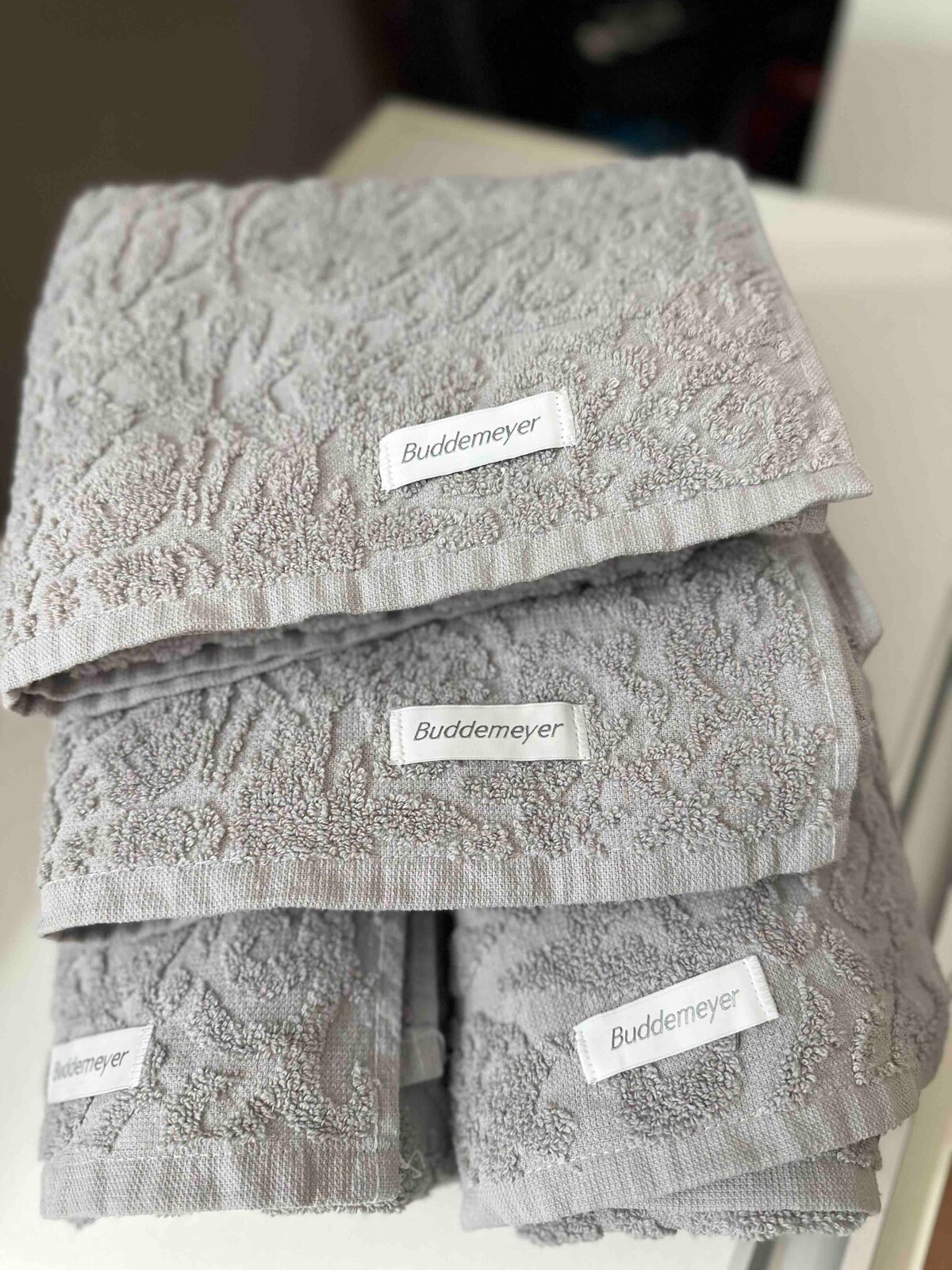 A neatly arranged stack of soft gray towels is displayed. Each towel features a decorative pattern and a label reading 'Buddemeyer,' indicating quality. The towels are folded neatly on a flat surface, showcasing their plush texture.