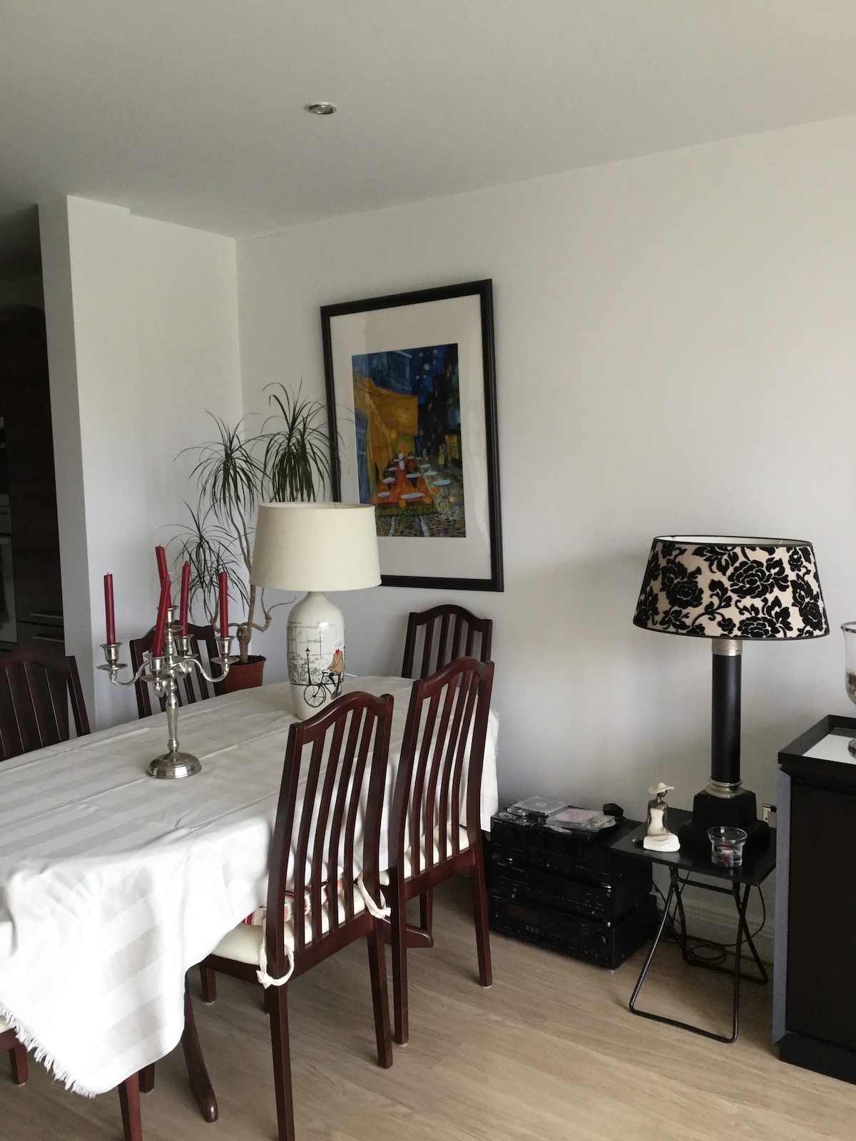 A dining area is depicted with a light-colored table set for six, surrounded by wooden chairs. A decorative lamp with a black and white pattern is positioned beside a tall plant. A colorful piece of artwork is hung on the wall, adding interest to the space.