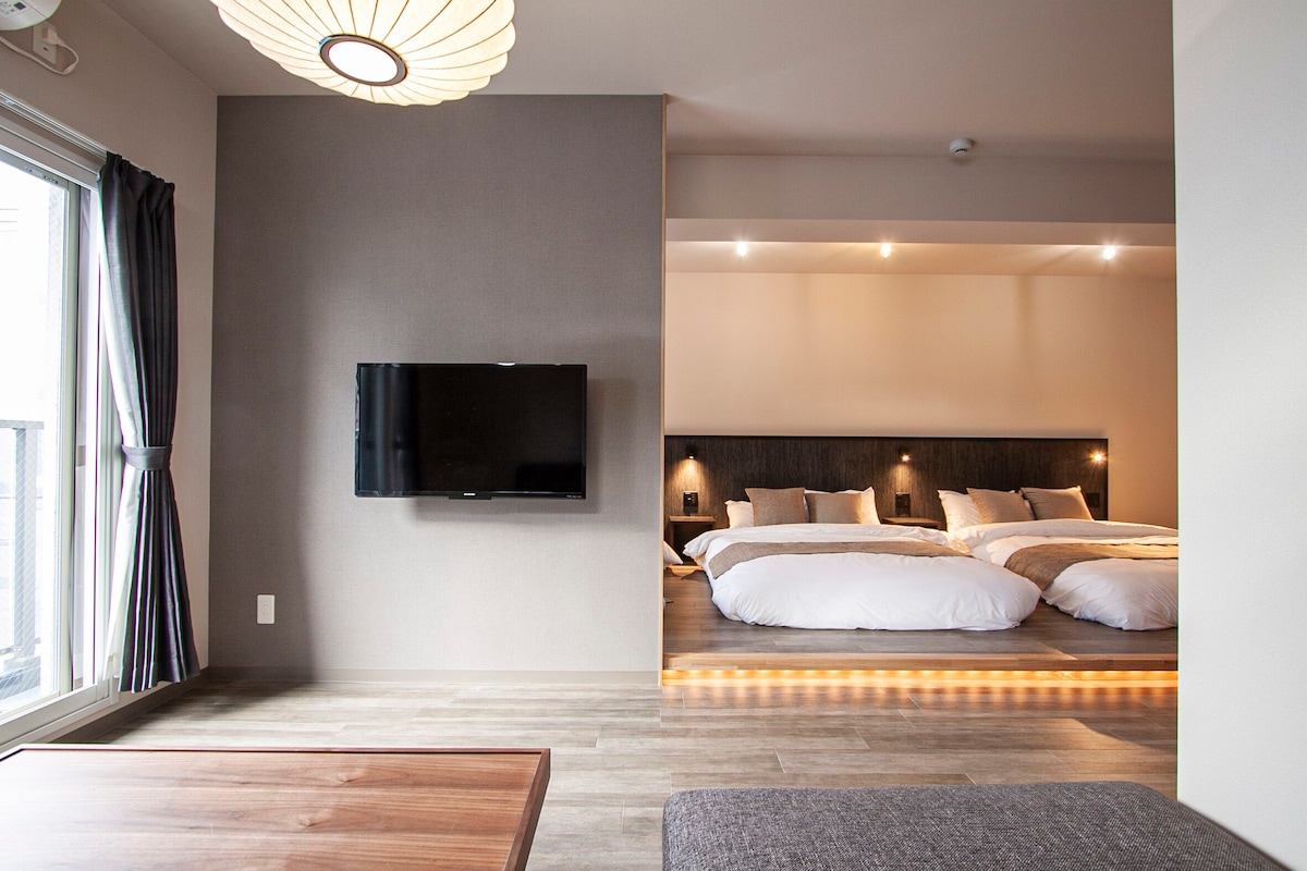 A spacious indoor area is presented, featuring a large window with curtains and a television mounted on the wall. The sleeping area contains multiple beds arranged in a row, adorned with neutral-toned bedding. A wooden table is positioned in the foreground, complementing the modern decor.