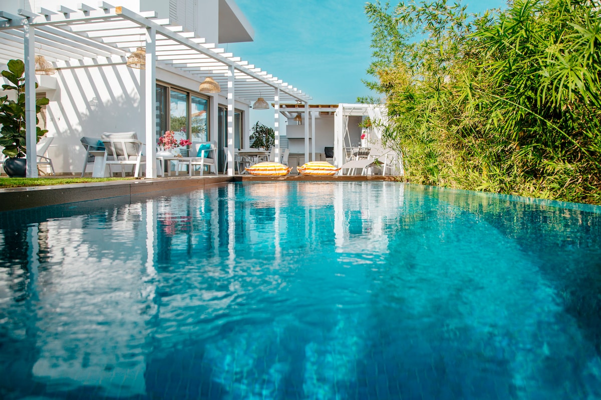 The private swimming pool features clear, shimmering water surrounded by a well-tended garden. Sunbeds are positioned poolside under the pergola, providing a comfortable space for relaxation. Lush greenery, including palm trees and bamboos, complements the serene outdoor setting.