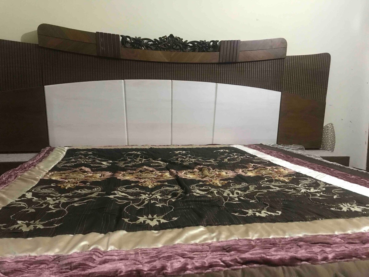 A spacious bed features an ornate headboard with intricate designs. A decorative bedspread with floral patterns in rich colors is layered on top, offering an elegant touch to the sleeping area.