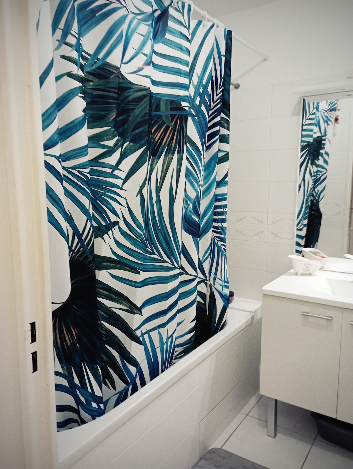 A clean bathroom features a shower curtain with a tropical leaf design, adding color to the white tiled space. A simple vanity with a mirror and sink is visible, complemented by a small arrangement of toiletries. Bright lighting enhances the overall ambience.