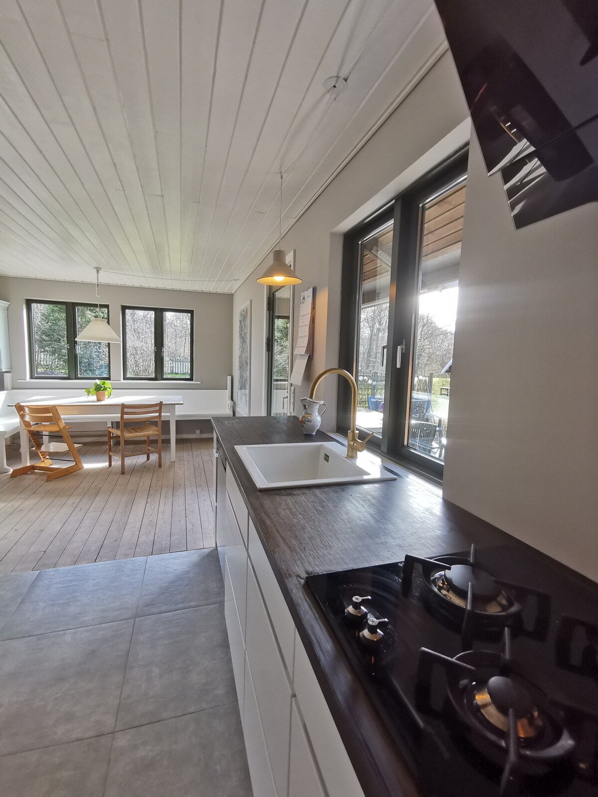 A modern kitchen area is presented with a sleek countertop and a built-in stovetop. Sunlight filters through large windows, illuminating wooden dining chairs and a table nearby. The minimalist design features light-colored cabinetry, promoting a spacious and functional feel.