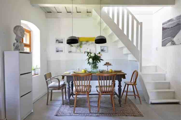 A dining area is presented with a wooden table surrounded by six chairs, featuring a floral centerpiece. Above, two black pendant lights hang from the ceiling. A staircase is visible in the background, accompanied by white walls adorned with framed photographs.