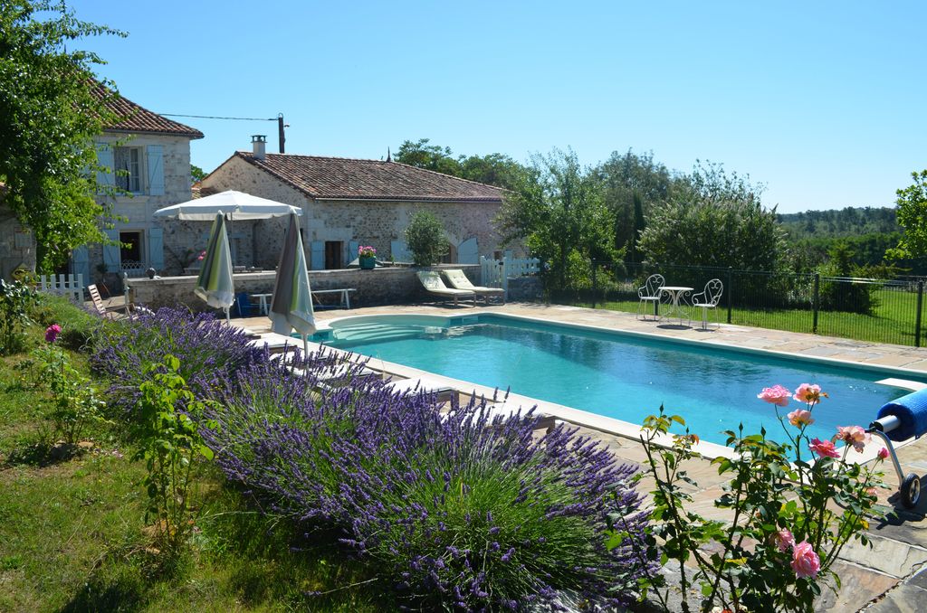 A large outdoor heated pool is framed by a sunlit stone patio and vibrant lavender flowers. Comfortable loungers are positioned around the pool, and a shaded seating area is available under a parasol. The picturesque stone farmhouse is visible in the background, surrounded by greenery.