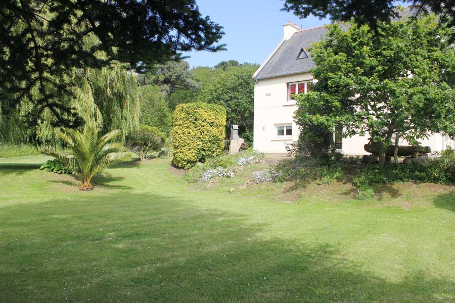 A spacious garden surrounds the house, featuring lush greenery and colorful flower beds. Large trees provide shade, while a palm tree adds a tropical touch. The exterior of the house displays a light-colored façade with red accents, merging harmoniously with the natural landscape.