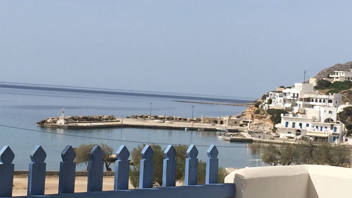 A calm seaside view is presented, showcasing a serene harbor with smooth water reflecting the sky. White buildings are observed on the hillside, complementing the coastal landscape. A wooden fence with blue accents offers a charming foreground to the tranquil scene.