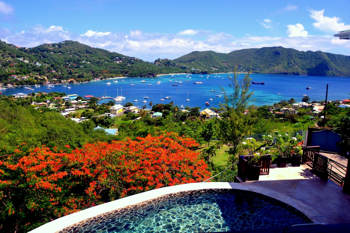 A panoramic view of Admiralty Bay is captured, showcasing the vibrant blue waters dotted with boats. Lush greenery and vibrant orange flowers frame the scene, while a sparkling private pool offers a relaxing spot to take in the stunning landscape.