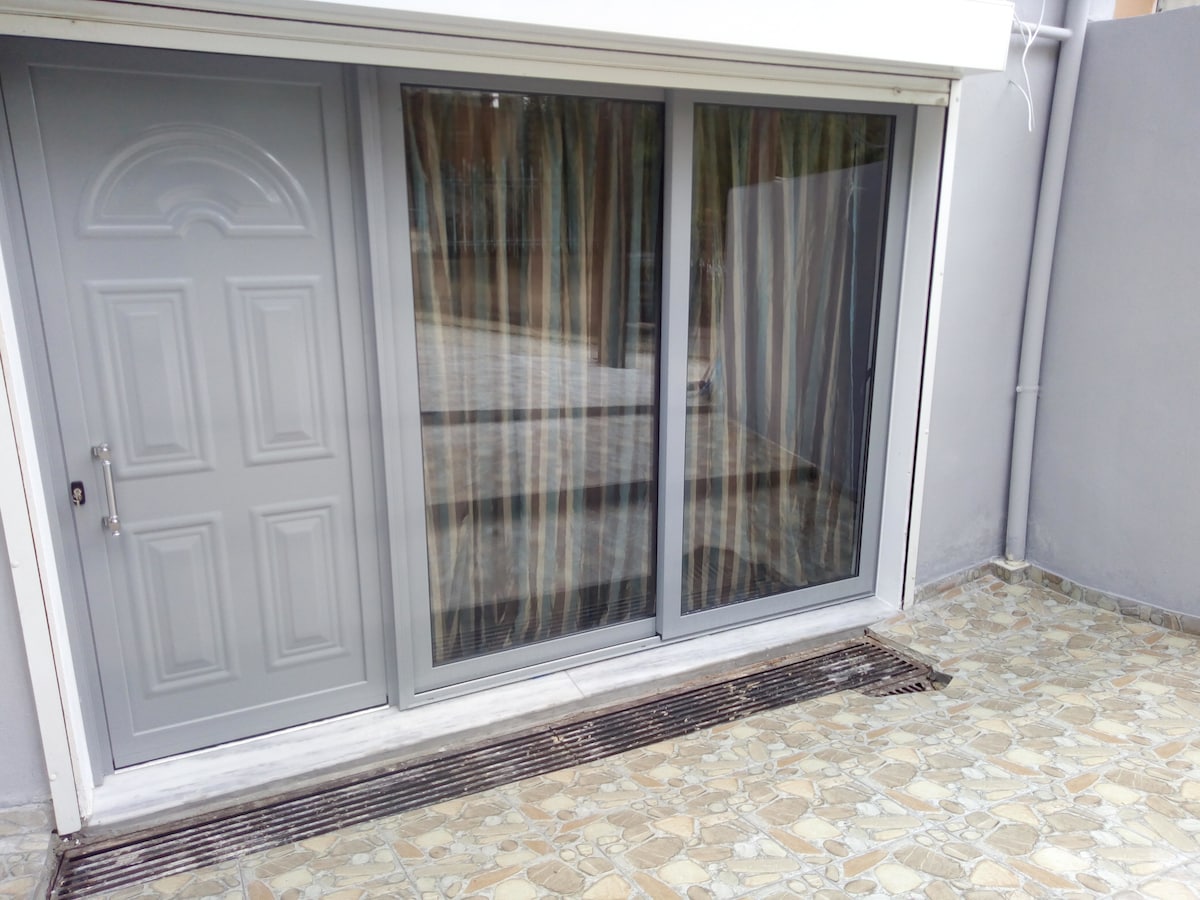 A sliding glass door is set within a gray wall, providing access to the outside. The door features sheer, patterned curtains that filter natural light. A stone pathway leads to the door, surrounded by a smooth, tiled area, creating a seamless transition between indoor and outdoor spaces.