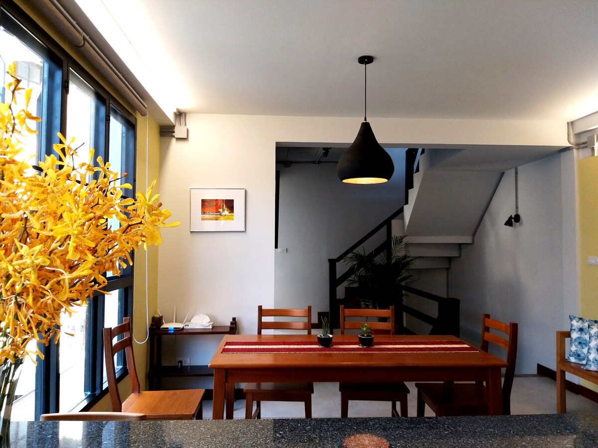 The dining area showcases a wooden table surrounded by six chairs, complemented by a black pendant light overhead. A vibrant arrangement of yellow flowers adds a touch of color. A staircase in the background leads to the upper floors, and a small desk is visible at the side.