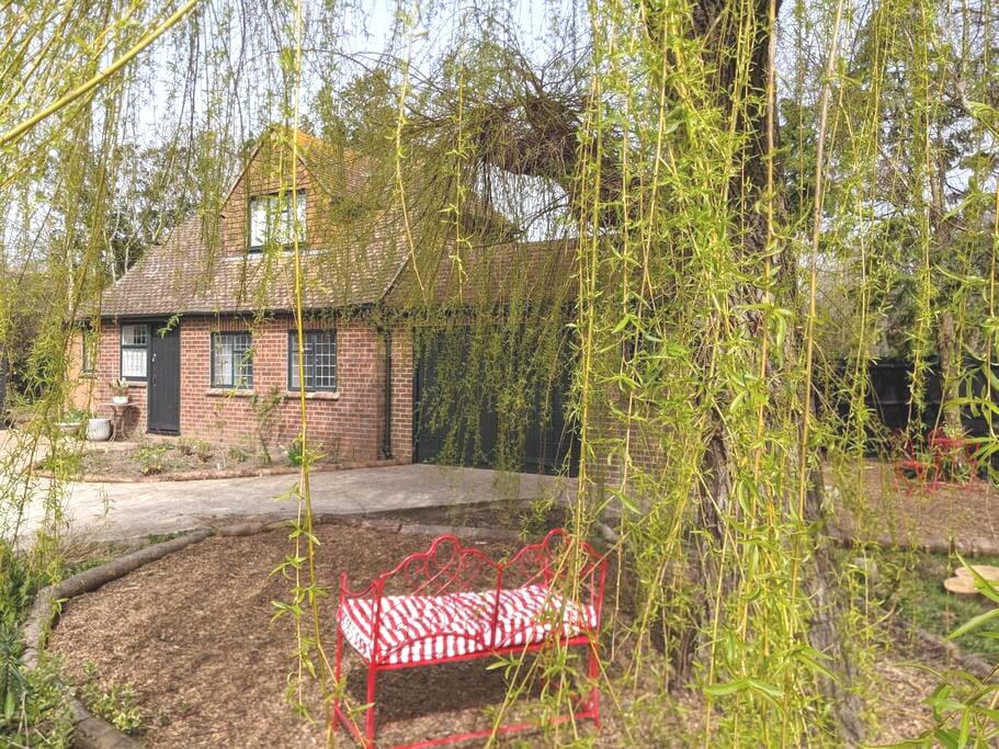 A charming red bench with white stripes is positioned under a cascading willow tree, providing a serene outdoor seating area. The brick cottage features a pitched roof and large windows, set against a backdrop of lush greenery and a gravel pathway.
