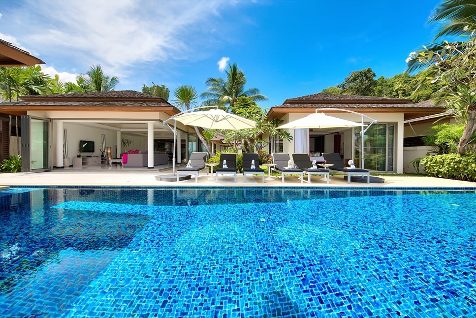 A large outdoor area features a shimmering infinity pool, surrounded by six sunbeds with umbrellas. Two open-air pavilions are visible in the background, complemented by lush tropical landscaping and palm trees under a clear blue sky.