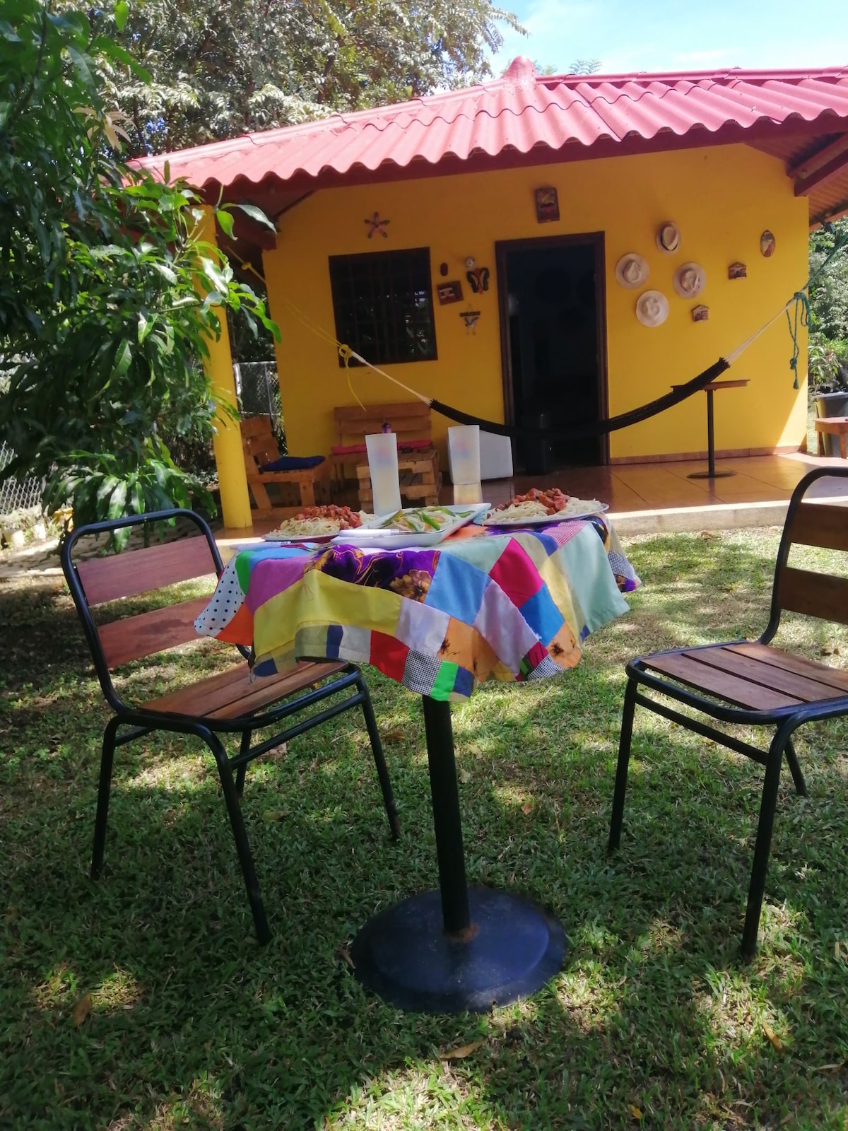 A colorful tablecloth covers a round table, set on a lush green lawn. Two chairs are positioned around the table, facing a bright yellow house with decorative plates on the exterior wall. The entrance is framed by lush greenery and a red roof.