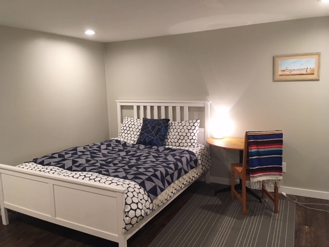 A guest room features a white bed adorned with a geometric patterned duvet, complemented by patterned pillows. A small wooden table is situated nearby, accompanied by a decorative throw on the chair. Soft lighting illuminates the space, enhancing the calm ambiance.