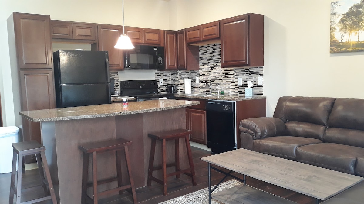 The kitchen area features rich wooden cabinetry and granite countertops, complemented by black appliances. A dining island with two stools is present, alongside a cozy seating area that includes a brown leather sofa and a wooden coffee table.