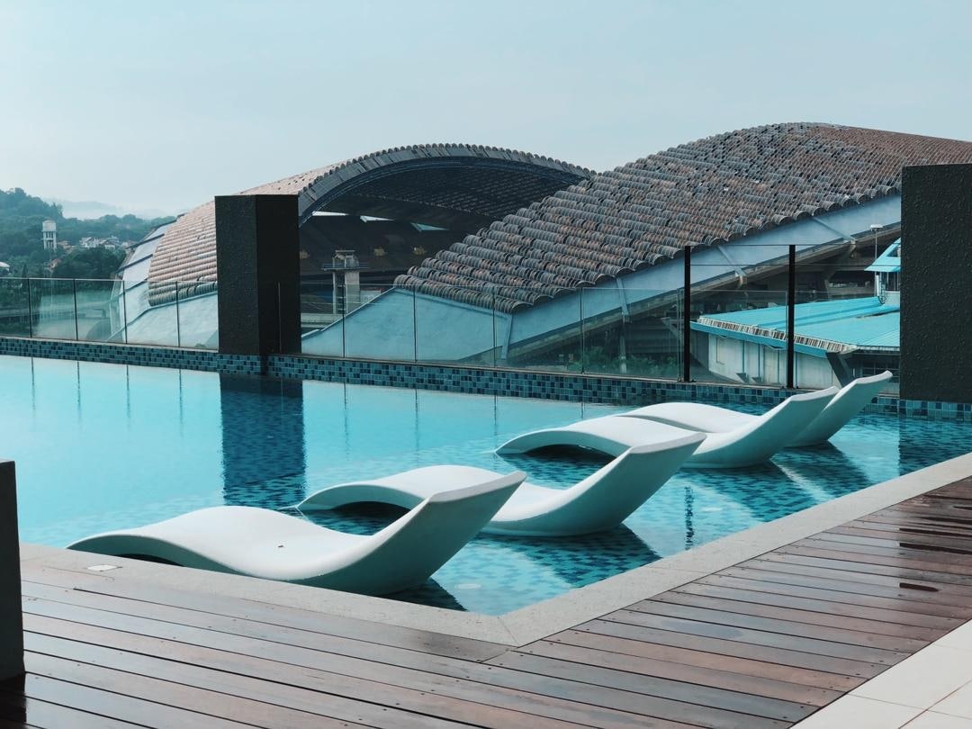 A serene pool area features four modern lounge chairs positioned along the edge of a shimmering blue pool. The backdrop includes a large, curved building with a textured roof, creating a contemporary and inviting environment.