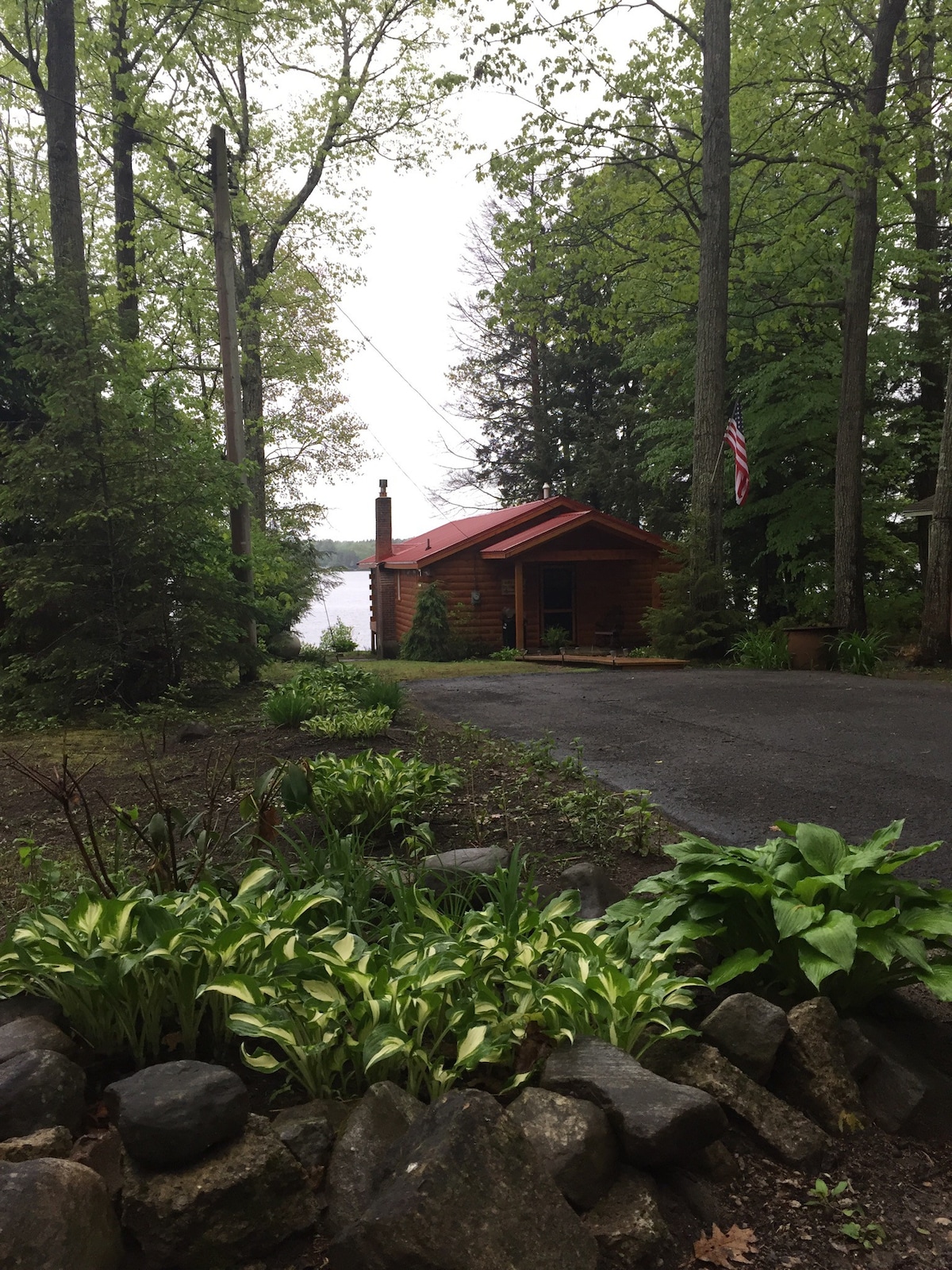 A cozy cabin with a red roof is surrounded by lush greenery, positioned along a gravel driveway. Natural stone landscaping frames the front garden, while nearby trees provide shade. A subtle view of the lake is visible in the background, enhancing the tranquil setting.