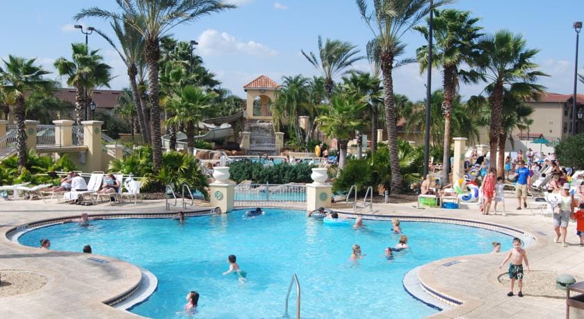 A large swimming pool is surrounded by tropical palm trees, with a variety of guests enjoying the water. Sun loungers are arranged around the pool area. In the background, additional resort amenities and lush landscaping are visible under a clear blue sky.