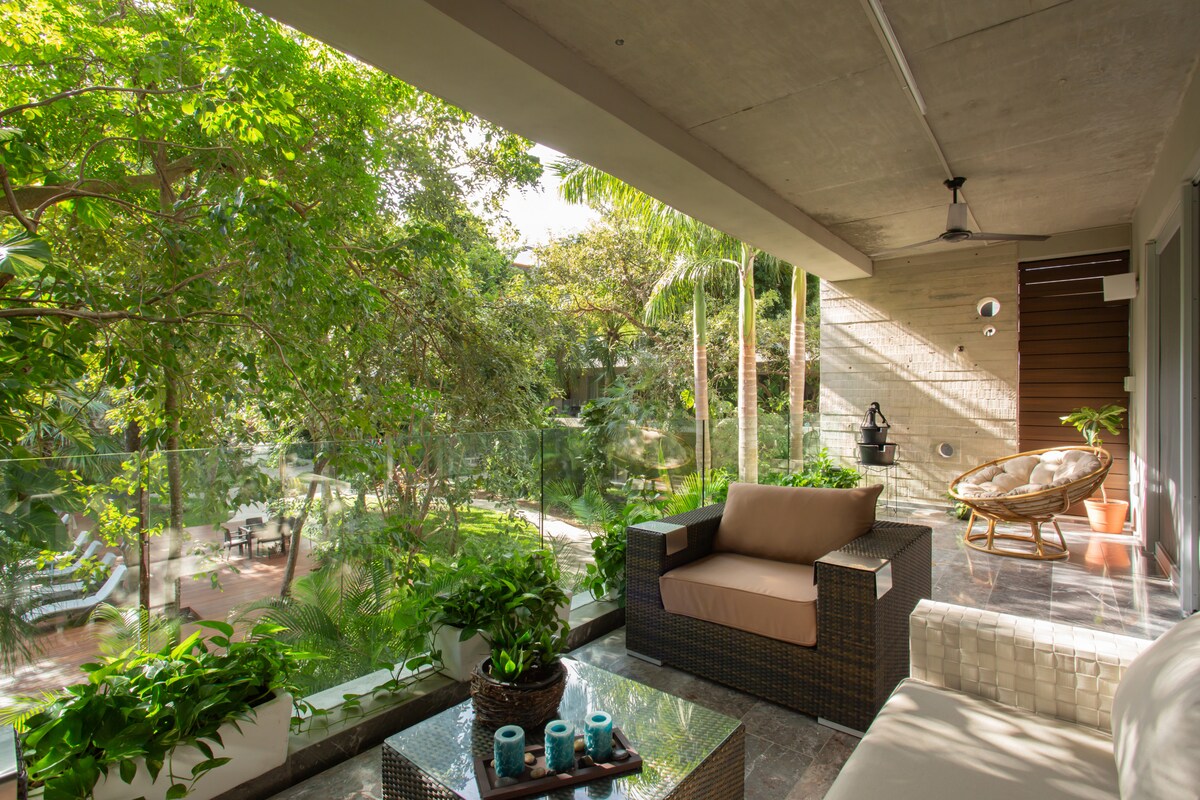 A shaded balcony area features a comfortable seating arrangement with a sofa and lounge chair, surrounded by lush greenery. A glass railing provides an unobstructed view of the landscaped garden below, where tropical plants and paths are visible.