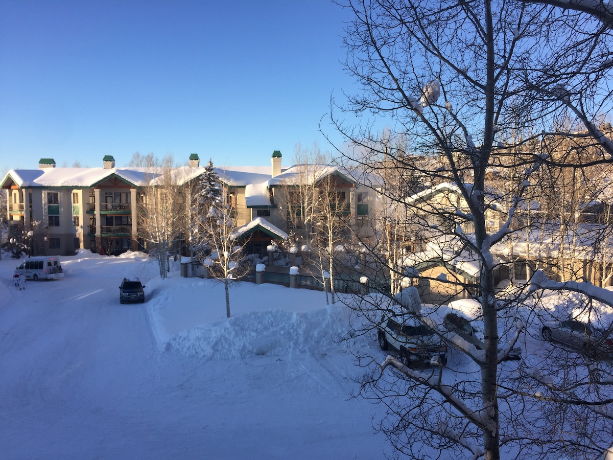 A winter scene outside the condo featuring a snow-covered parking area. Several vehicles are parked among snowdrifts, with a backdrop of the building complex partially obscured by trees. Clear blue skies are visible above, indicating crisp winter weather.