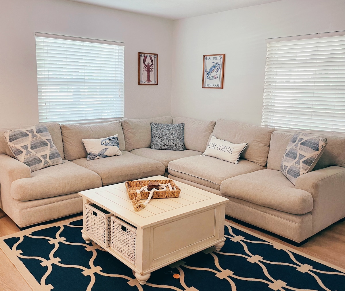 A spacious living area features a large sectional sofa adorned with various decorative pillows. A light-colored coffee table is positioned in the center, complemented by a decorative basket. Natural light filters through two window blinds, creating a bright and inviting environment.