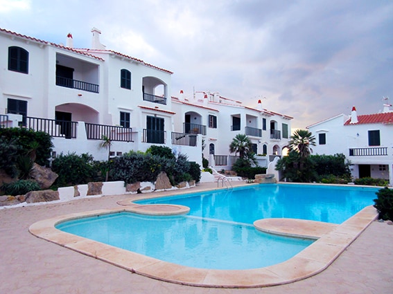 A bright swimming pool is surrounded by a sandy area and lush greenery, with apartments featuring white facades and balconies in the background. Sun loungers are placed along the water's edge, and a few palm trees provide a tropical feel.