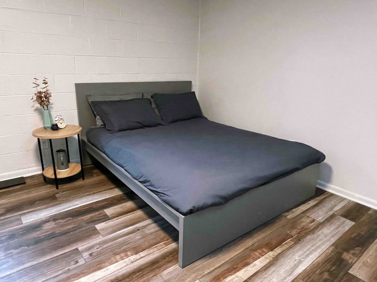 A cozy sleeping area features a spacious bed dressed in dark bedding. A circular side table with a small decorative element is positioned beside the bed. The room has light-colored walls and a wood-like floor, creating a warm and inviting environment.