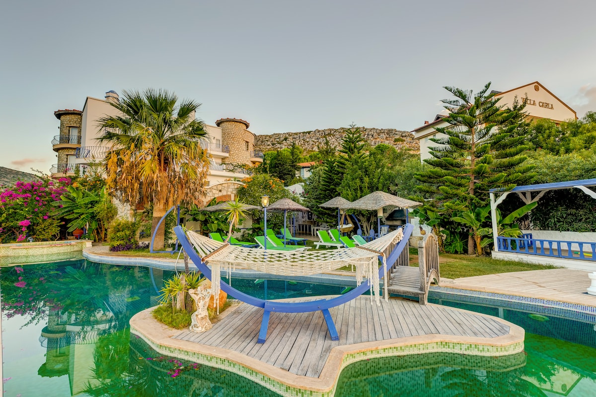A tranquil pool area features a blue wooden hammock strung between two posts, surrounded by palm trees and colorful plants. Lounge chairs in green are positioned nearby. The water reflects the lush greenery and the architectural details of the building in the background.
