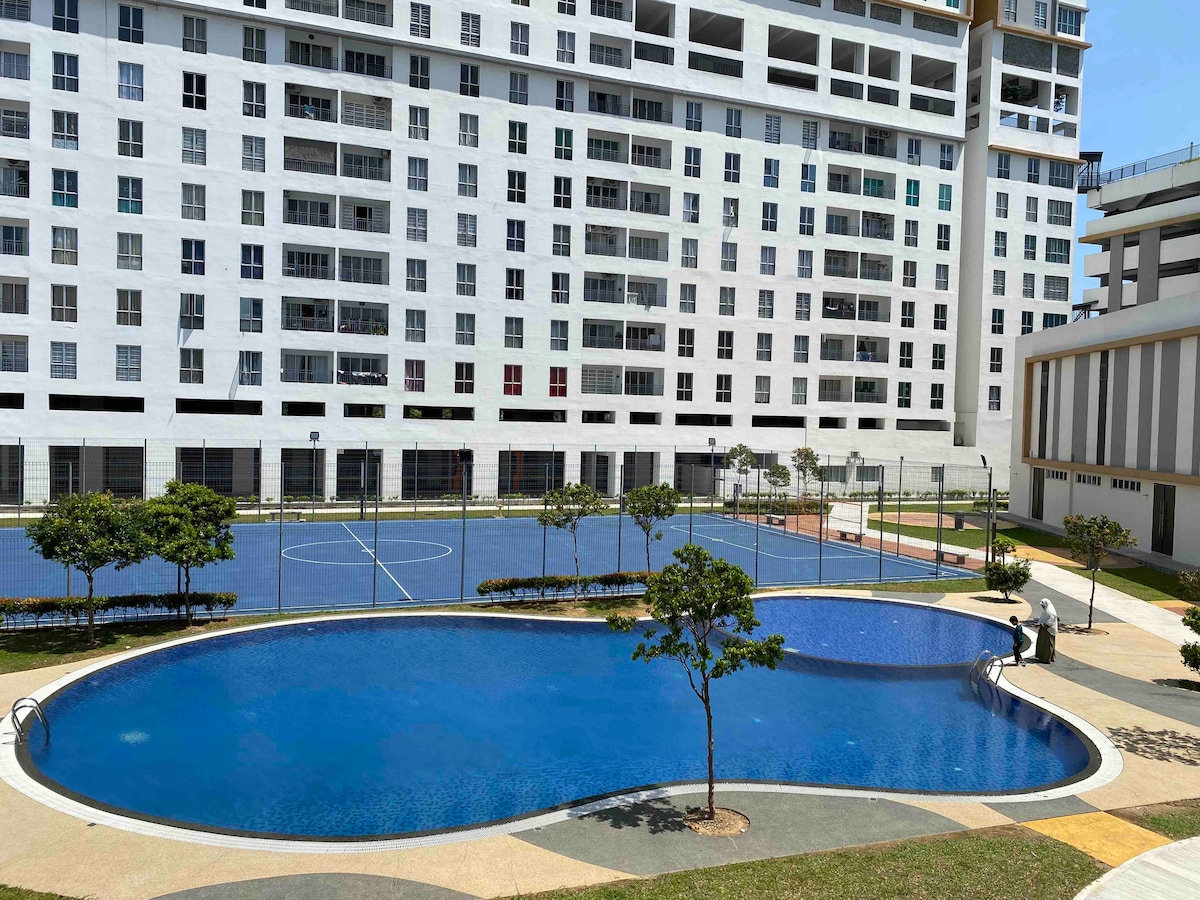 A large swimming pool is surrounded by manicured landscaping, featuring several trees. Adjacent to the pool, a spacious outdoor area includes sports courts. The modern building structure provides a backdrop, showcasing a variety of windows that reflect sunlight.