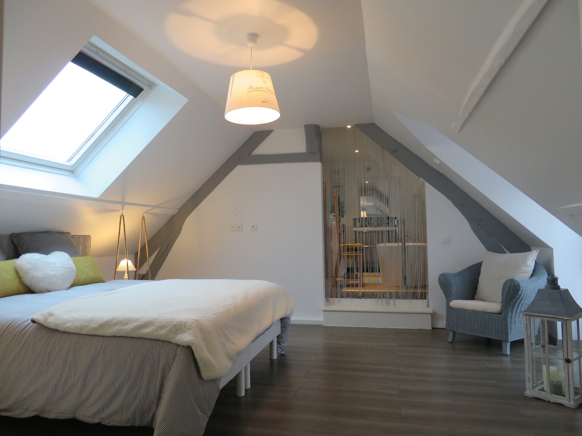 A bright attic bedroom features a spacious bed adorned with soft bedding. Natural light pours in from a skylight, illuminating the minimalist decor. A cozy armchair and decor elements add character, while a divider subtly separates the sleeping area from a small nook in the background.